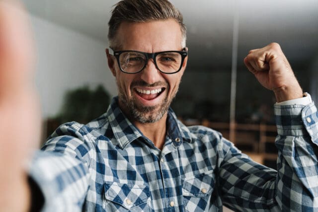 Excited white-haired man making winner gesture while taking selfie photo